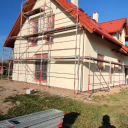 Dom jednorodzinny w trakcie malowania elewacji, otoczony rusztowaniem na tle błękitnego nieba, z widocznym stosem podestów rusztowania na trawie i cieniem osoby.