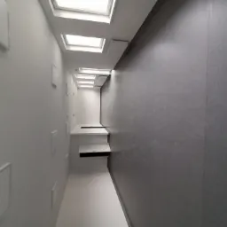 Długi korytarz z białymi ścianami z wnękami i szarą ścianą, oświetlony szeregiem okien dachowych.