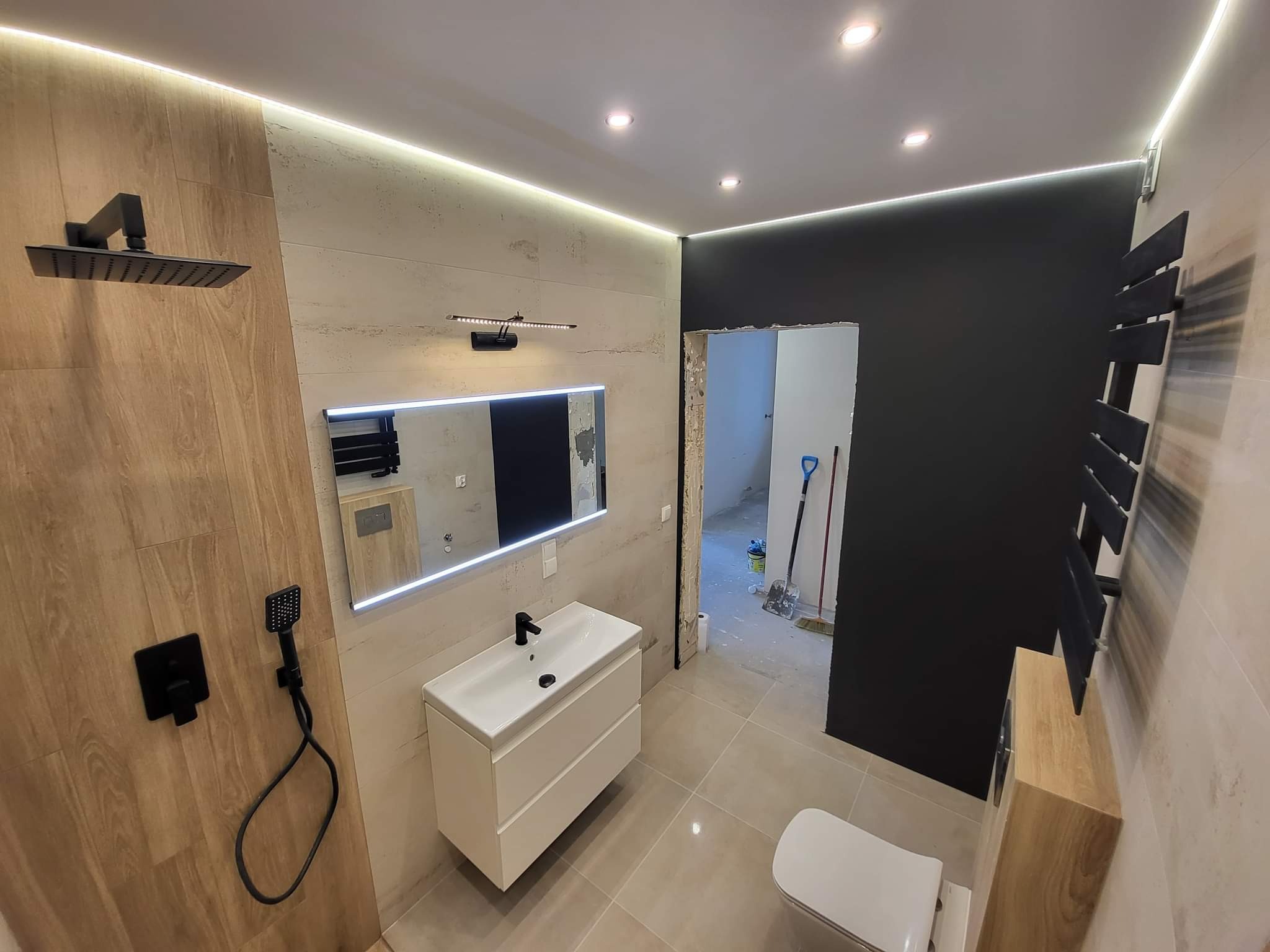 Nowoczesna łazienka z beżowymi płytkami imitującymi drewno, czarną armaturą i podświetlanym lustrem, widoczne niedokończone prace w sąsiednim pomieszczeniu.