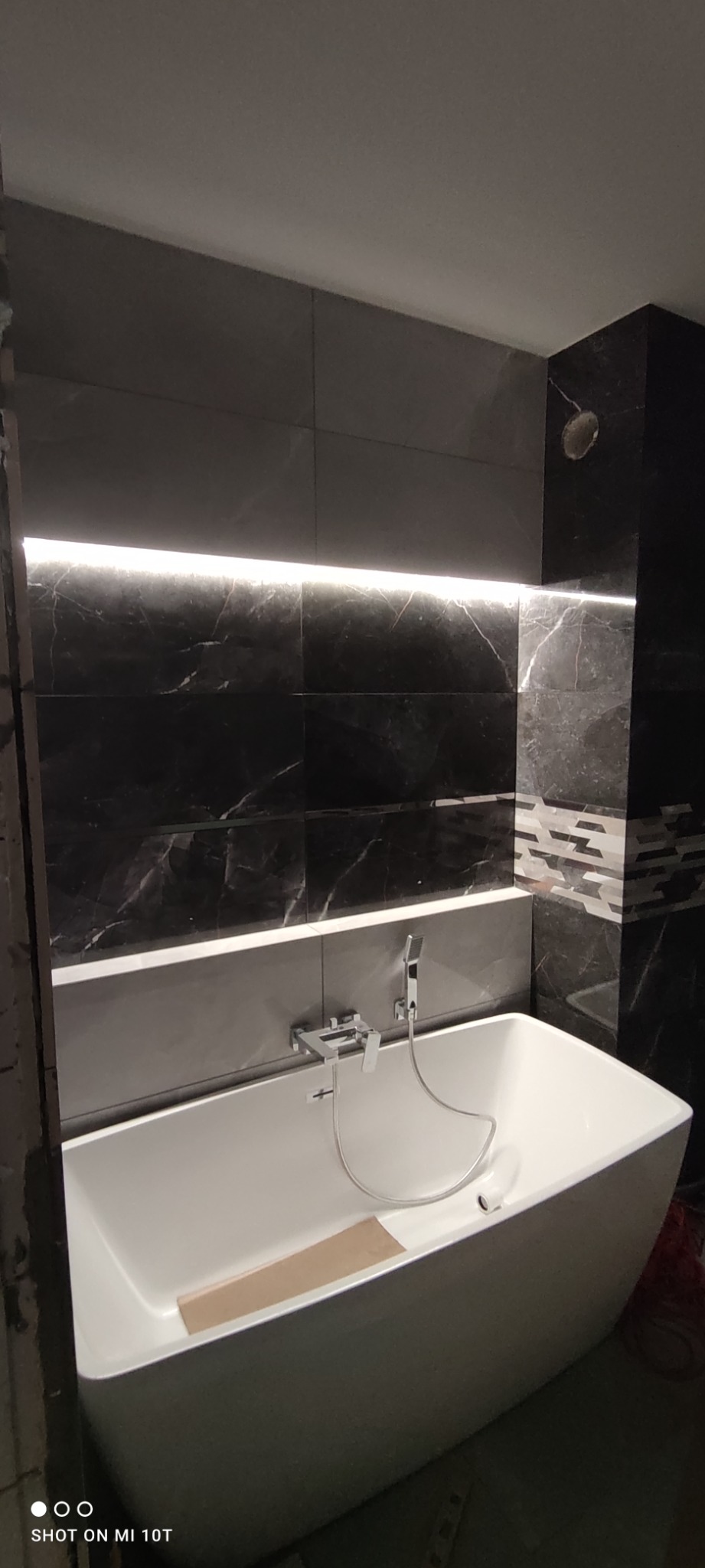 Nowoczesna łazienka z czarnymi marmurkowymi płytkami, podświetleniem LED nad wanną i białą, wolnostojącą wanną z chromowaną armaturą. Na dnie wanny leży karton.