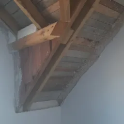 Skośny sufit poddasza z drewnianą konstrukcją nośną i fragmentami obróbki tynkarskiej przy ścianie, widok z dołu.