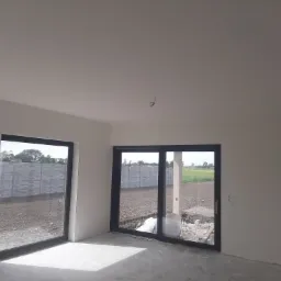 Wnętrze nowo wykończonego pokoju z dwiema przesuwnymi szklanymi drzwiami balkonowymi, widok na zewnątrz na plac budowy i pole, gładkie białe ściany i sufit, surowa podłoga.