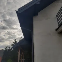 Fragment elewacji domu z nowym, ciemnoszarym orynnowaniem i podbitką dachową, widoczne ślady po naprawach na elewacji, pochmurne niebo w tle.