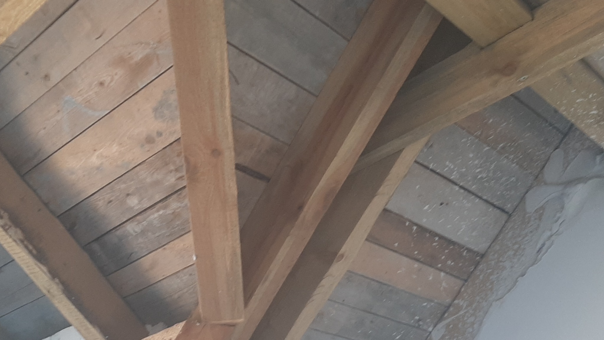 Skośny widok na drewniany szkielet poddasza z desek, z widocznymi elementami konstrukcyjnymi i fragmentami ściany z pozostałościami zaprawy.