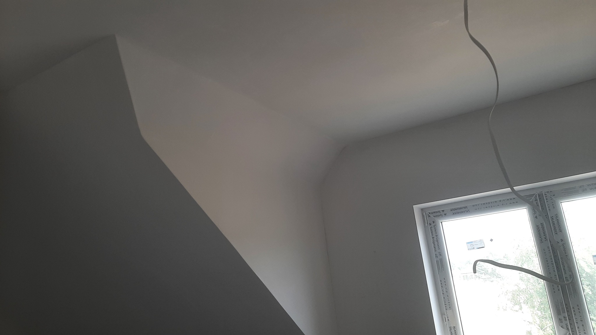 Wykończone wnętrze poddasza ze skosem, pomalowane na biało, widoczne okno PCV z naklejkami producenta i wiszący kabel elektryczny.