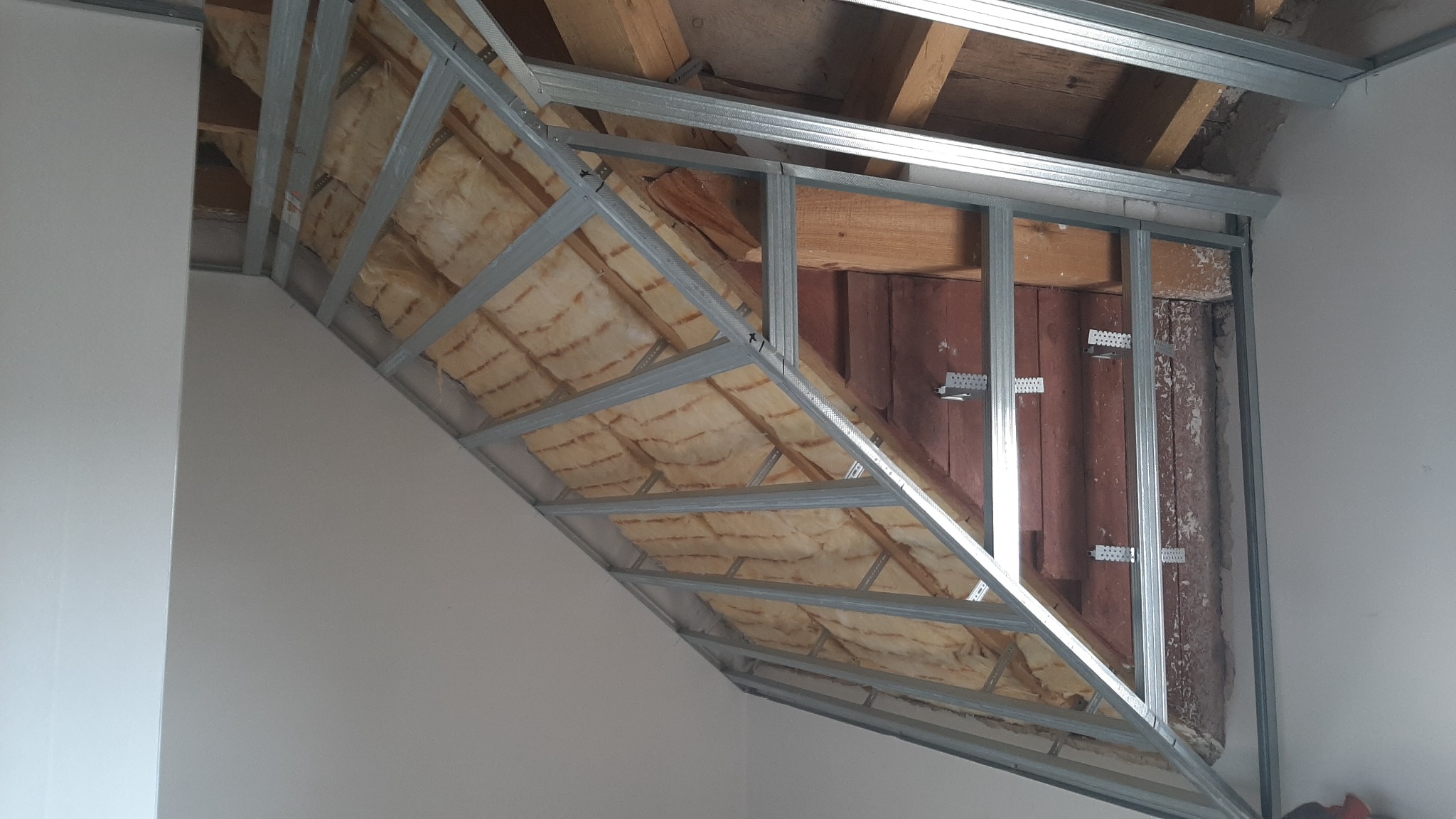 Skośna konstrukcja metalowa poddasza z izolacją z wełny mineralnej, widoczne elementy drewniane stropu i ściany, przygotowanie do montażu okładziny.