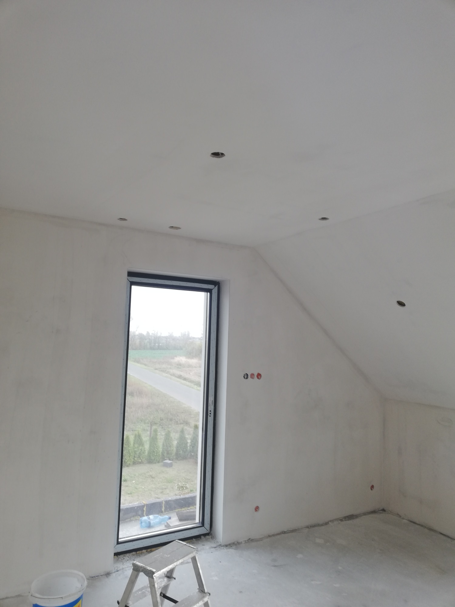 Poddasze po szpachlowaniu i przygotowaniu pod malowanie, widoczne otwory na oświetlenie punktowe w suficie, okno z widokiem na zewnątrz, drabina i wiadro farby na podłodze.