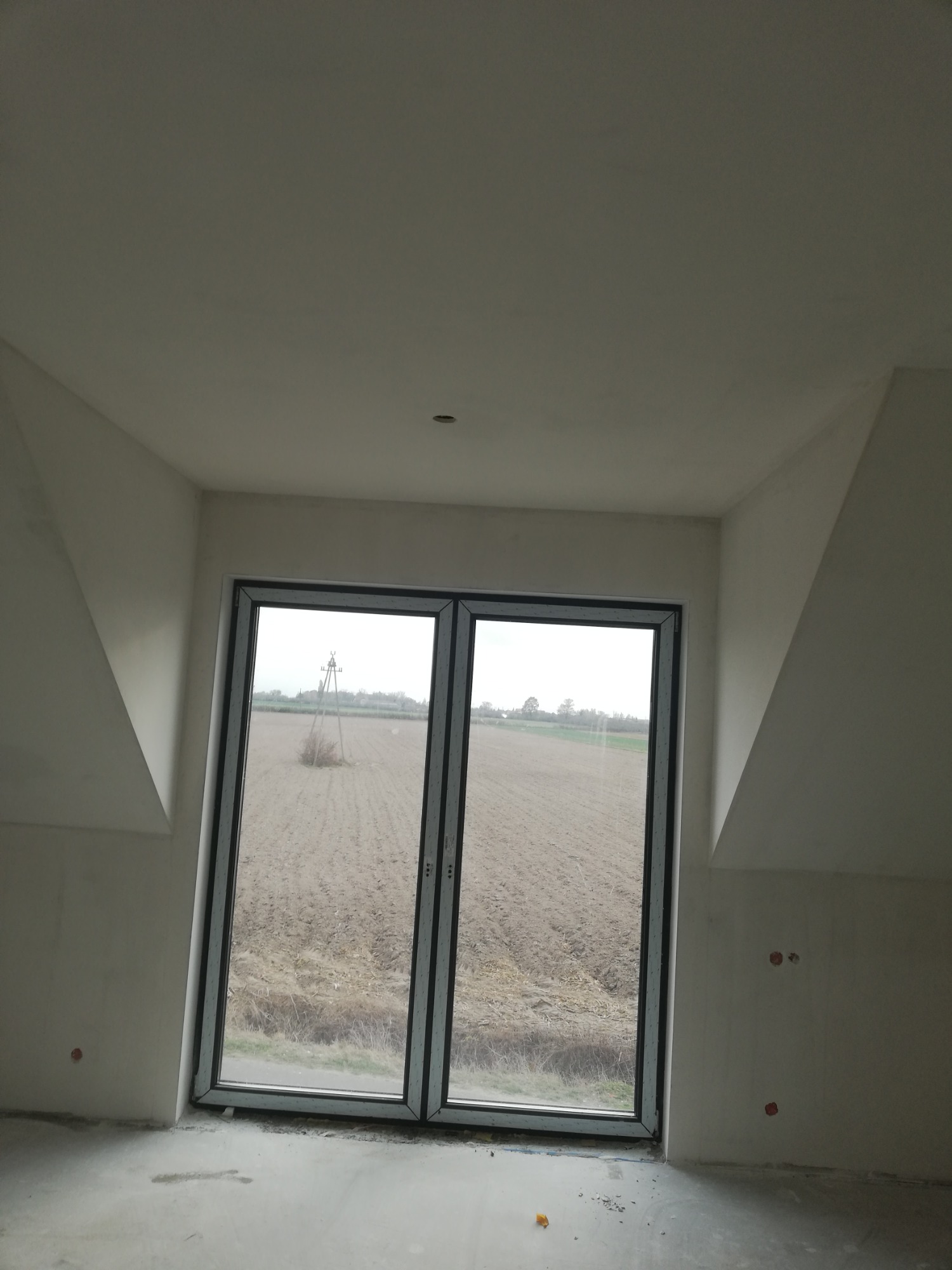 Wnętrze poddasza z oknem balkonowym, ściany przygotowane pod malowanie, widoczne niedoskonałości szpachlowania i otwory po gniazdkach elektrycznych.