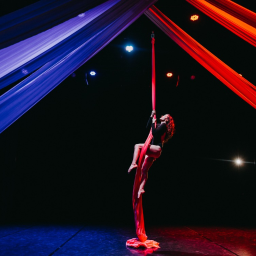 Zakład Pracy Kreatywnej - Kobieta wykonuje akrobacje na czerwonej szarfie, oświetlona reflektorami, z niebieskimi i czerwonymi tkaninami w tle.