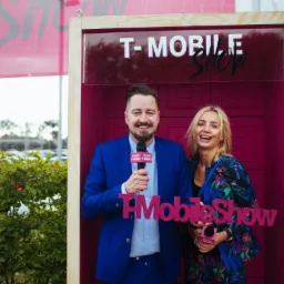 Dwoje uśmiechniętych prezenterów w studio plenerowym T-Mobile Show, mężczyzna w niebieskim garniturze trzyma mikrofon, kobieta w kwiecistej marynarce prezentuje logo, za nimi parking i zieleń.