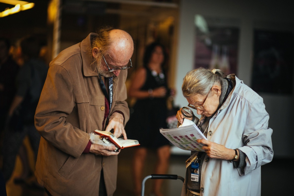 Dwie osoby starsze, mężczyzna z książką i kobieta z lupą, analizują dokumenty w zatłoczonym pomieszczeniu.