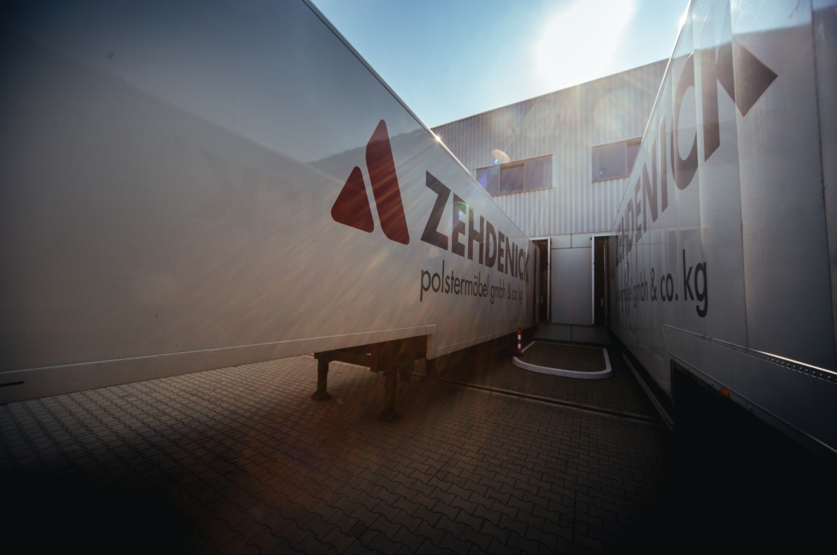 Dwa białe samochody dostawcze z logo firmy meblowej Zehdenick, stojące obok siebie na brukowanym placu w słoneczny dzień.