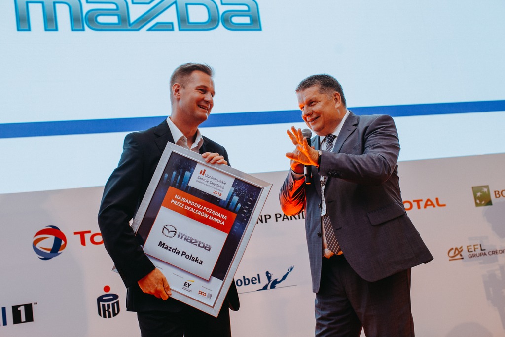 Dwóch mężczyzn w garniturach na scenie podczas wręczania nagrody dla Mazda Polska, na tle logo Mazda i sponsorów.