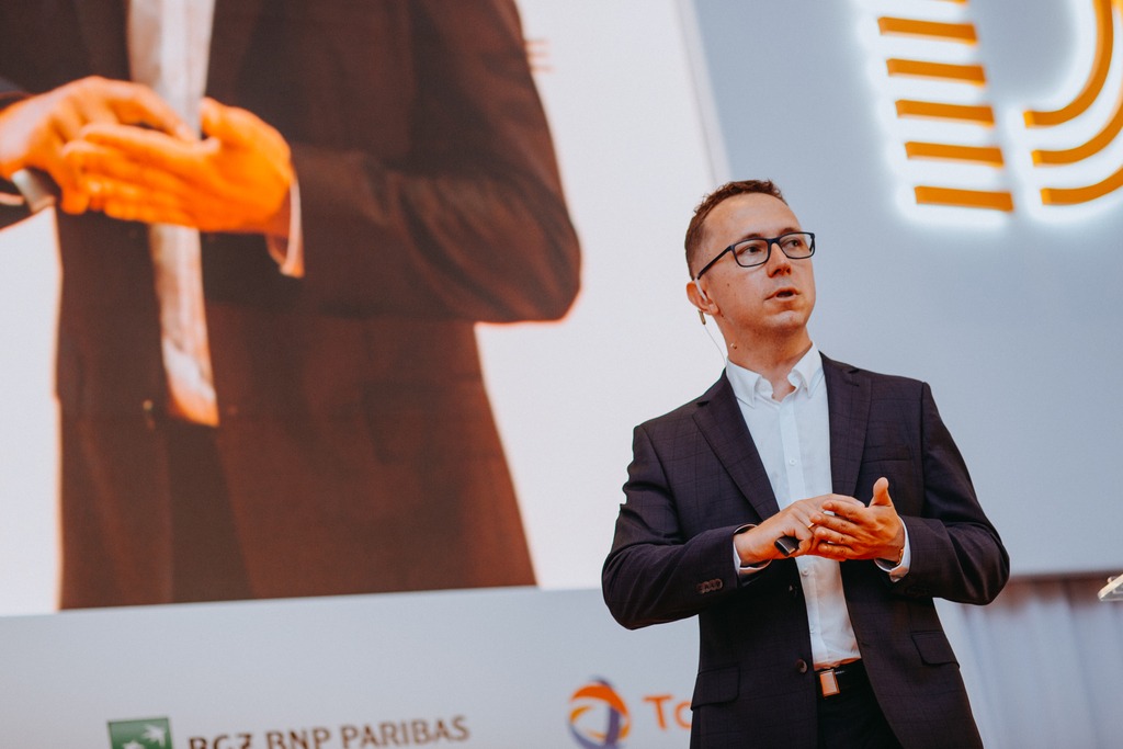 Mężczyzna w okularach i ciemnej marynarce przemawia na tle ekranu z logotypami firm, trzymając w dłoni pilot prezentacyjny.