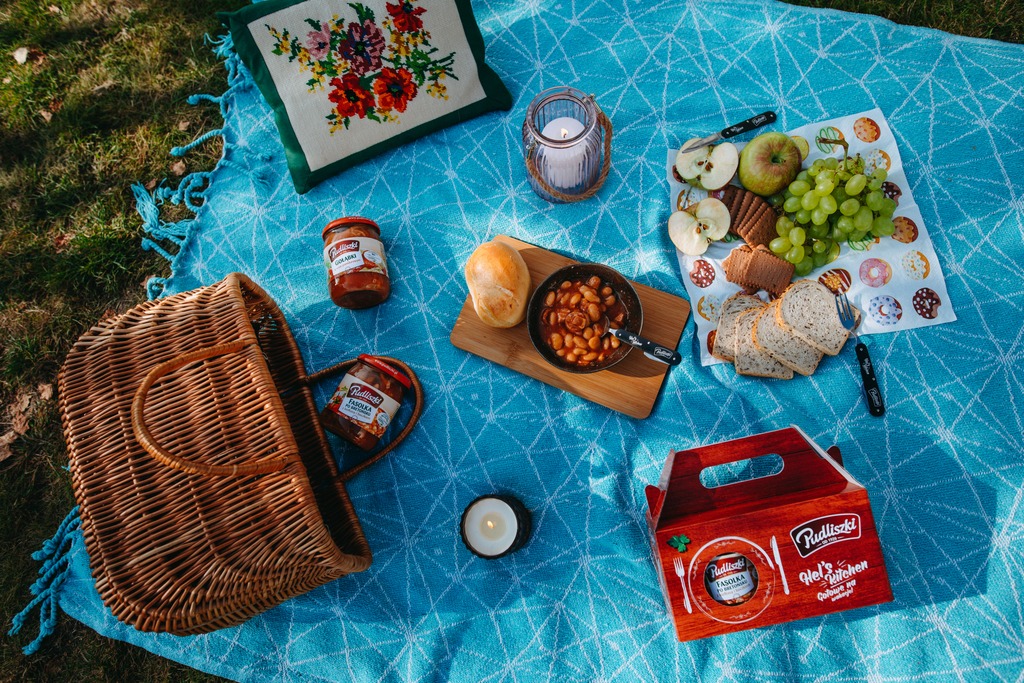 Rozłożony na trawie błękitny koc piknikowy z geometrycznym wzorem, na którym znajdują się: wiklinowy kosz, słoiki z przetworami, danie z fasoli w patelni, pieczywo, owoce, ciastka, świece...