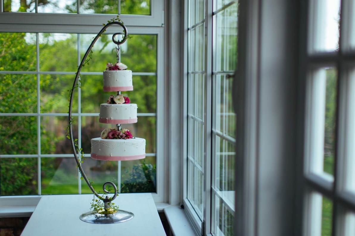 Trzypiętrowy tort weselny, biały z różowymi akcentami i kwiatami, zawieszony na metalowym stojaku z dekoracją z bluszczu, ustawiony na białym stole przy oknie z widokiem na zieleń.