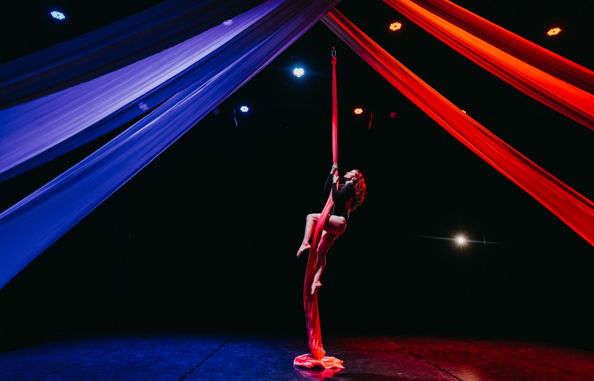 Kobieta wykonuje akrobacje na czerwonej szarfie, oświetlona reflektorami, z niebieskimi i czerwonymi tkaninami w tle.