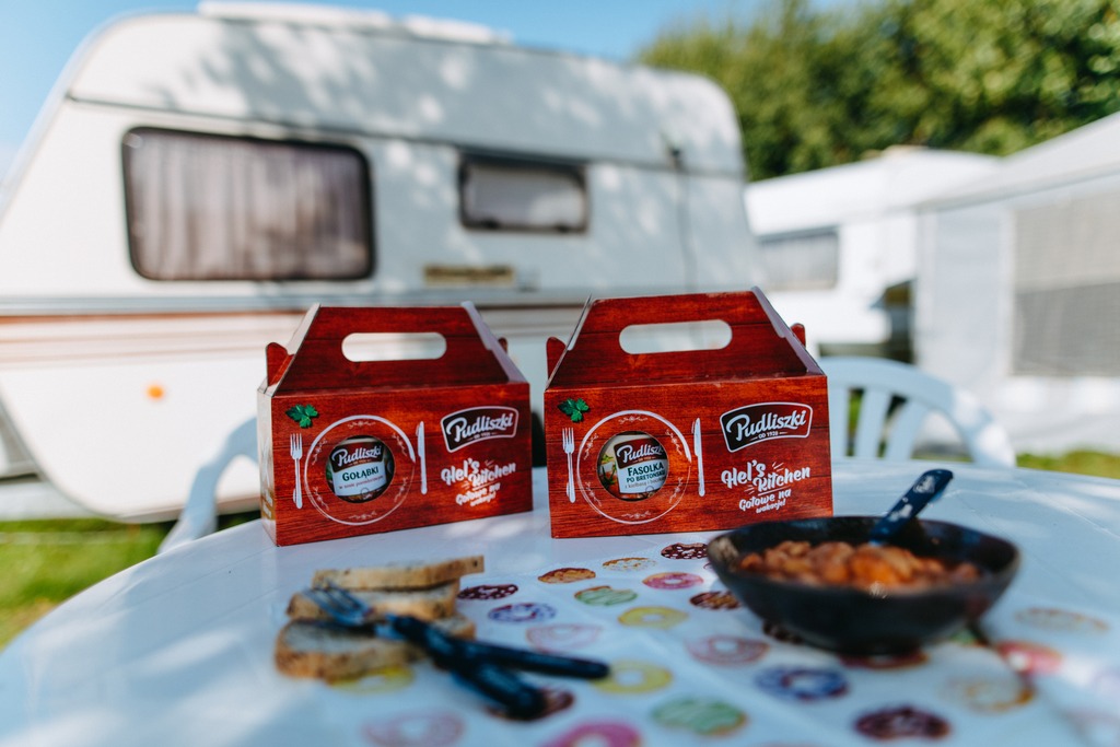 Dwa pudełka z gotowymi daniami firmy Pudliszki (Gołąbki i Fasolka po bretońsku) stoją na stole z nadrukiem pączków, obok miseczka z fasolką, chleb i sztućce, a w tle przyczepa kempingowa.