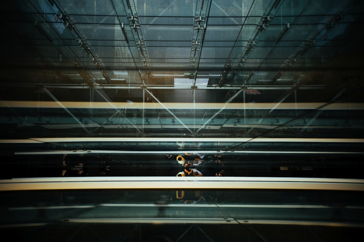 Widok z dołu na szklany sufit budynku biurowego z widocznymi elementami konstrukcyjnymi i odbijającymi się sylwetkami ludzi.