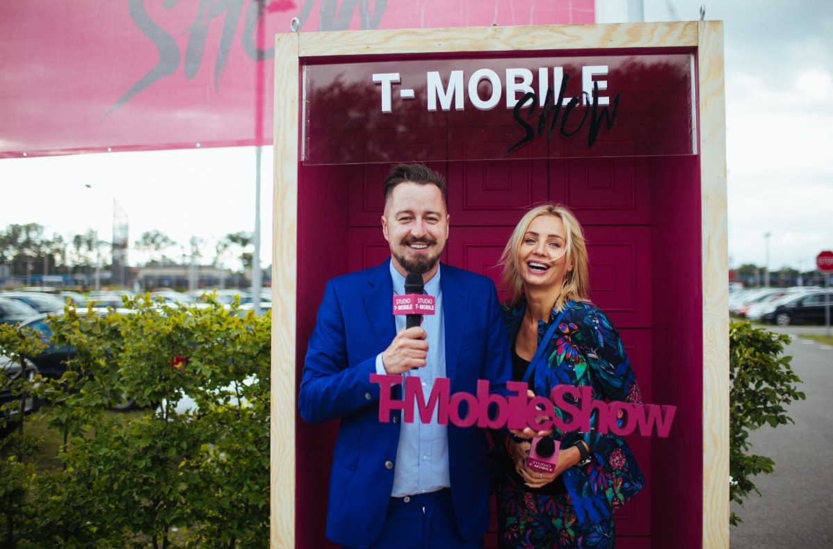 Dwoje uśmiechniętych prezenterów w studio plenerowym T-Mobile Show, mężczyzna w niebieskim garniturze trzyma mikrofon, kobieta w kwiecistej marynarce prezentuje logo, za nimi parking i zieleń.