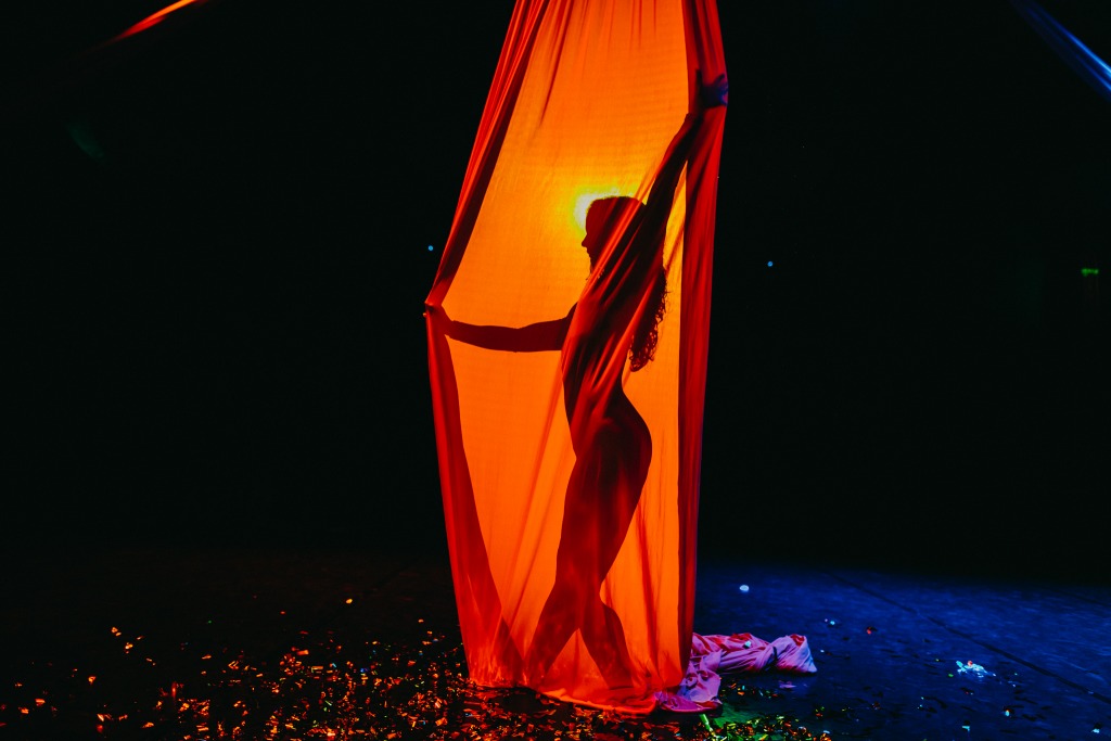 Sylwetka kobiety za pomarańczową tkaniną na scenie, oświetlona od tyłu, z konfetti na podłodze.