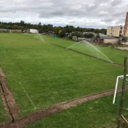 Rozległe boisko piłkarskie w trakcie instalacji automatycznego nawadniania, widoczne wykopane rowy i tryskacze w akcji, w tle budynki mieszkalne i pochmurne niebo.