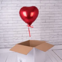 Czerwony balon w kształcie serca unosi się z białego pudełka na tle ściany z cegły.