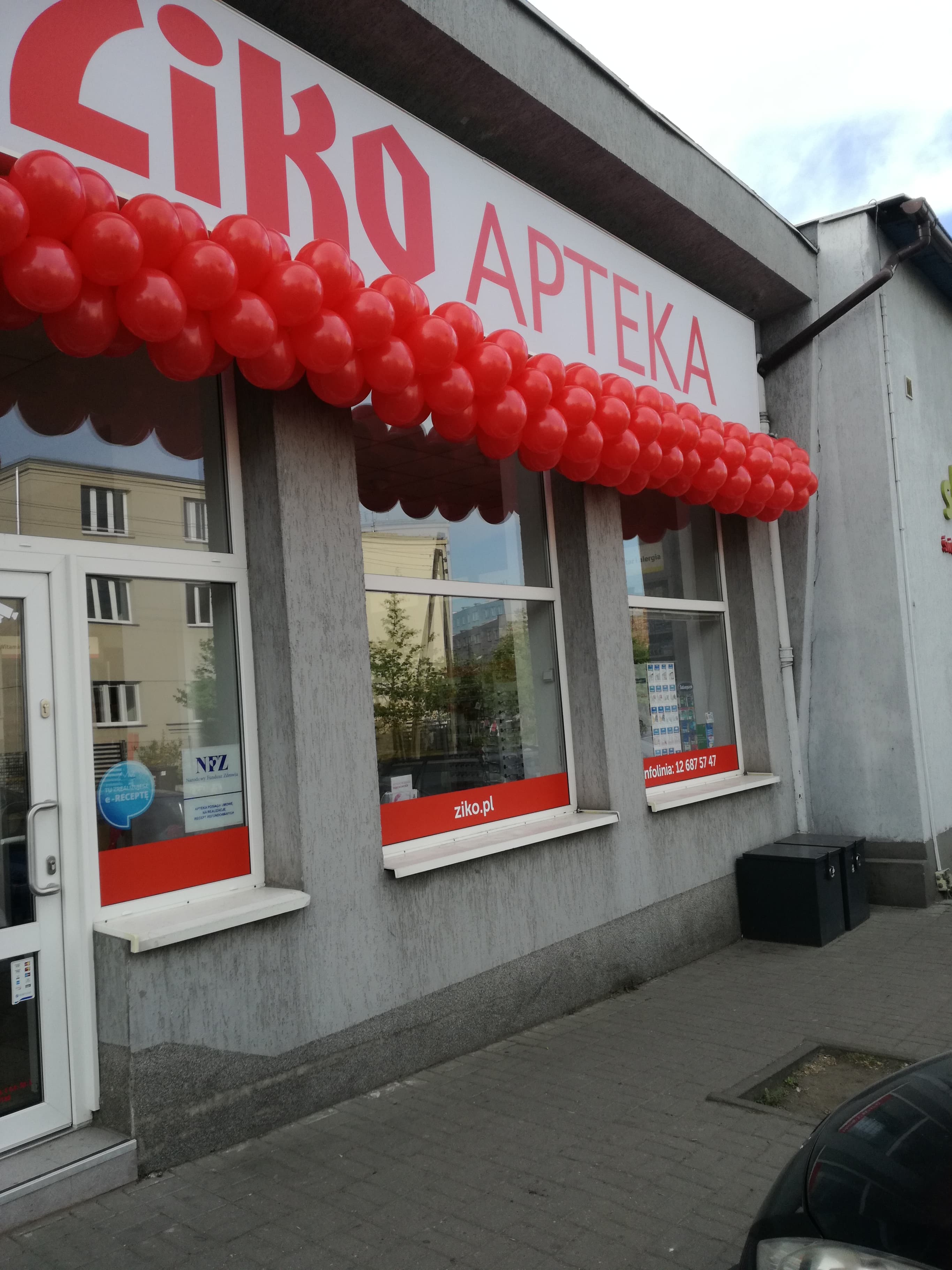 Fasada apteki udekorowana girlandą z czerwonych balonów, szyld z nazwą apteki widoczny powyżej okien i drzwi.