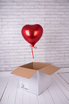 Czerwony balon w kształcie serca unosi się z białego pudełka na tle ściany z cegły.