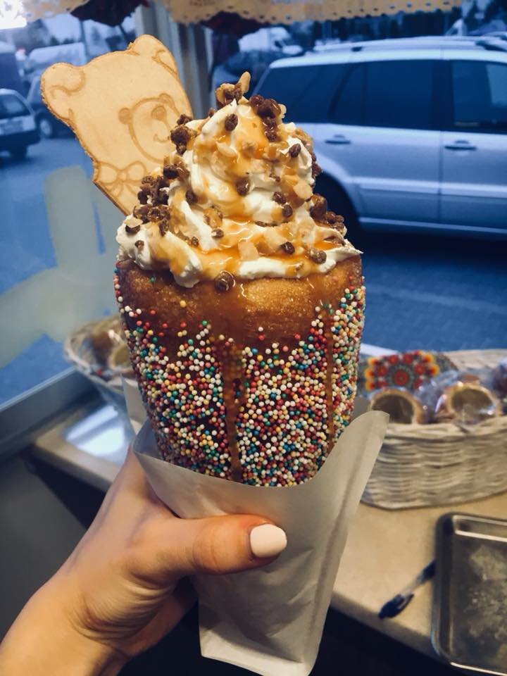 Trdelnik w papierowej owijce, obsypany kolorową posypką, z bitą śmietaną, karmelem i czekoladowymi kawałkami, udekorowany drewnianym misiem, trzymany w dłoni na tle okna i zaparkowanego samochodu.