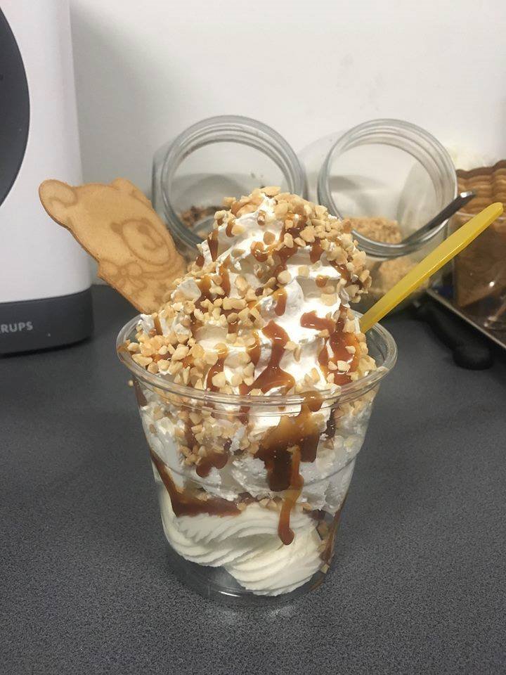 Deser lodowy w przezroczystym kubku, z bitą śmietaną, sosem karmelowym, posypką z orzechów i ciastkiem w kształcie misia, podany z żółtą łyżeczką, na szarym blacie, w tle słoiki z zawartością.