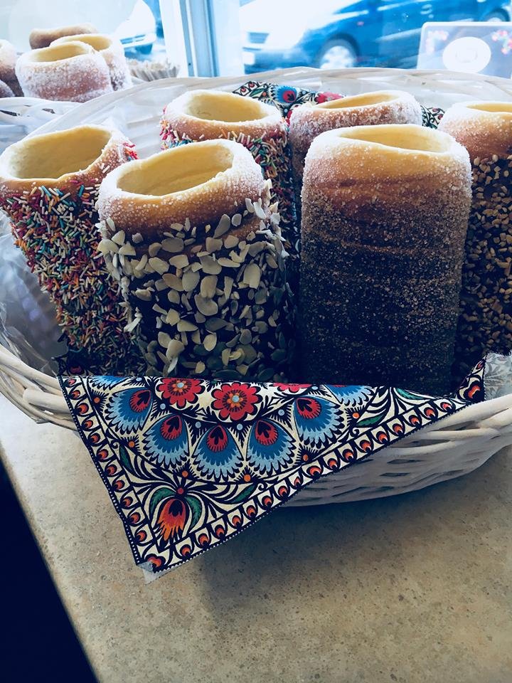 Kosz wiklinowy z tradycyjnymi węgierskimi ciastami kominkowymi (kurtoskalacs) posypanymi cukrem, orzechami, posypką i czekoladą, wyłożony chustą z ludowym wzorem.