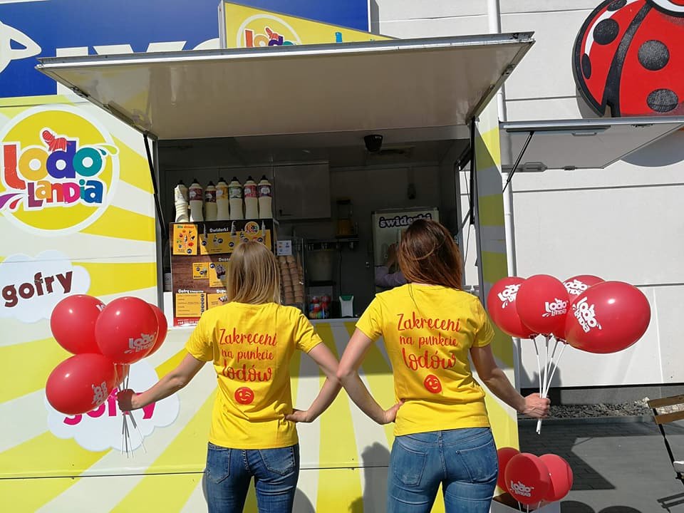 Dwie kobiety w żółtych koszulkach z napisem 'Zakręceni na punkcie lodów' stoją przed budką z lodami 'Lodo Landia', trzymając czerwone balony z logo. W tle widoczne menu i pojemniki z polewami.