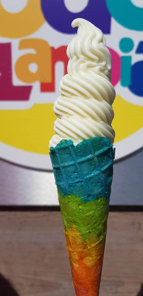 Wysoki, kremowy lód w wafelku barwionym w odcieniach niebieskiego, zielonego i pomarańczowego, na tle kolorowego logo.