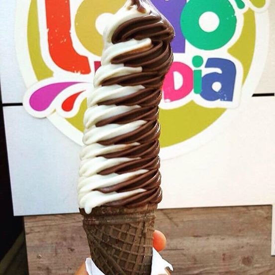 Wysoki, spiralny lód czekoladowo-waniliowy w ciemnym waflu, trzymany w dłoni, na tle kolorowego logo.