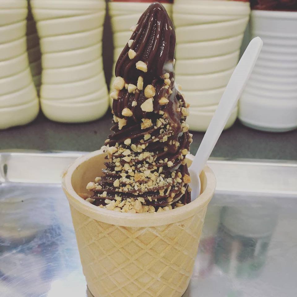 Czekoladowy deser lodowy w waflu, posypany orzechami, z białą łyżeczką, na metalowej tacy, z pojemnikami na lody w tle.