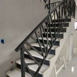 Nowoczesne schody z czarną, geometryczną balustradą, białe stopnie z czarnymi akcentami, widoczne instalacje elektryczne w ścianie, biała drabina malarska w tle.