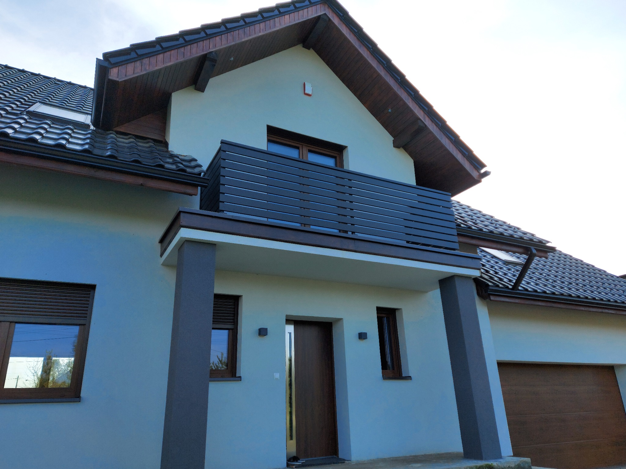 Nowoczesny dom jednorodzinny z balkonem zabezpieczonym poziomą balustradą z ciemnego drewna, widok z dołu na elewację z drewnianymi oknami i drzwiami wejściowymi.