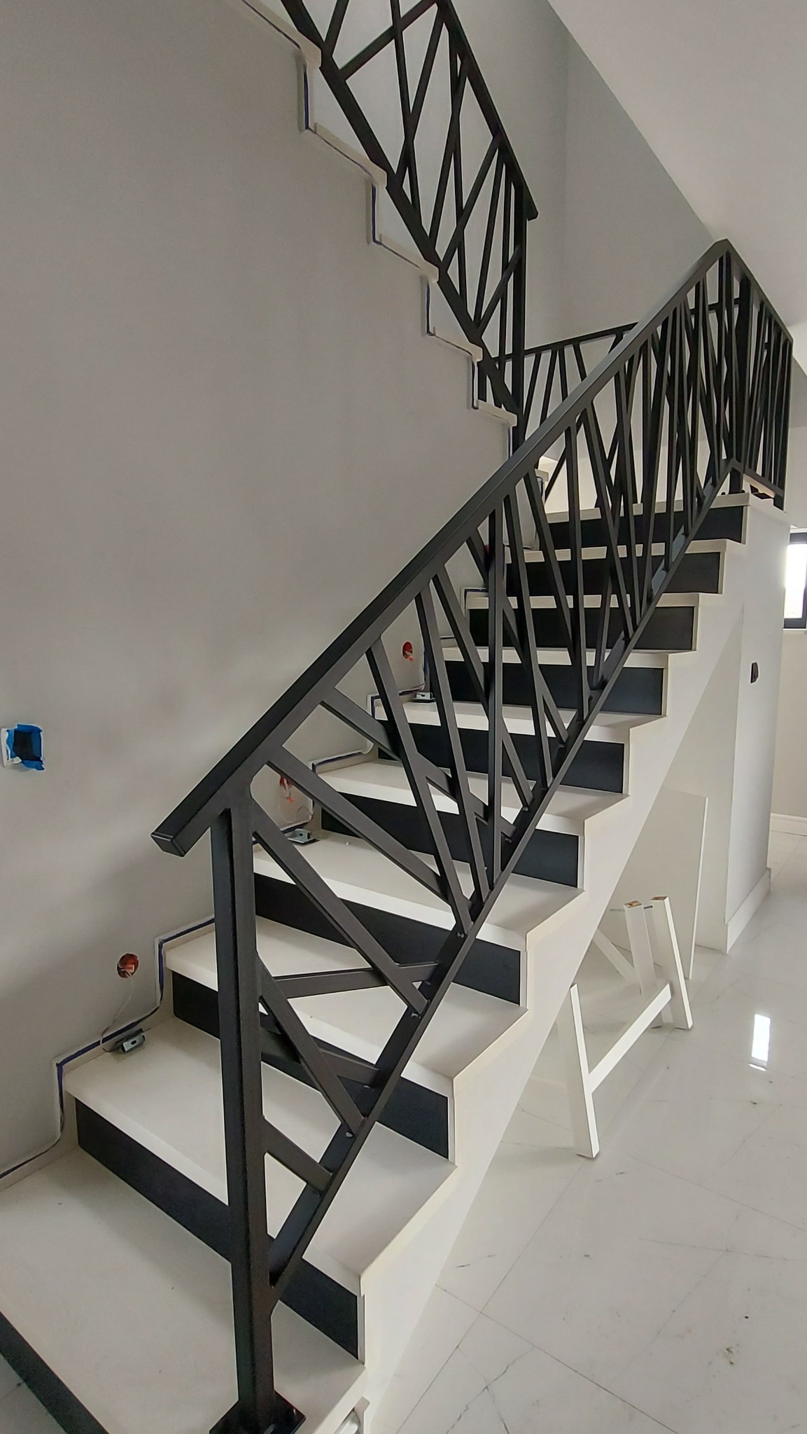 Nowoczesne schody z czarną, geometryczną balustradą, białe stopnie z czarnymi akcentami, widoczne instalacje elektryczne w ścianie, biała drabina malarska w tle.