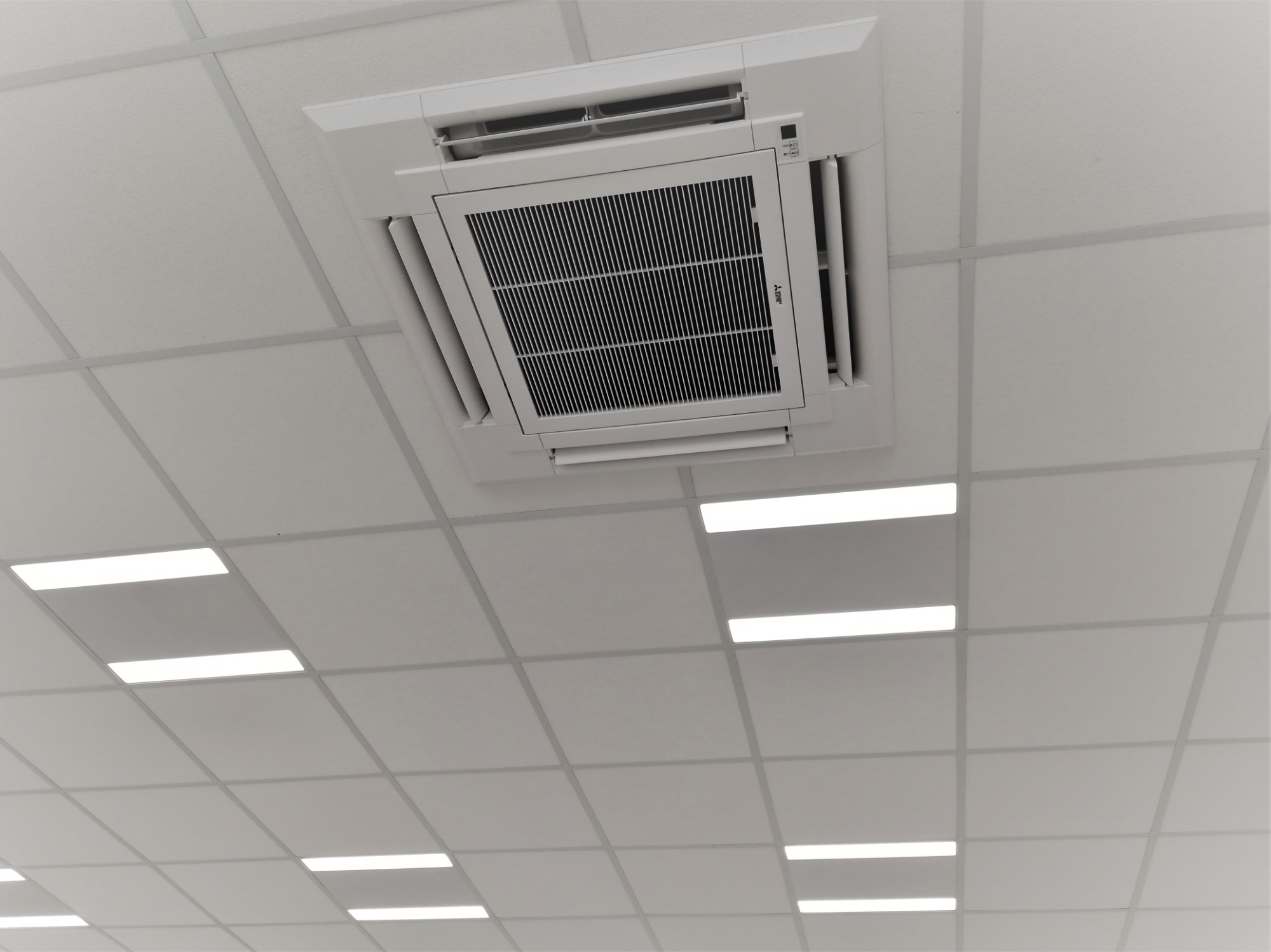 Kasetonowa biała sufitowa klimatyzacja marki Mitsubishi, widoczna z dołu, z kratką wlotową powietrza i regulowanymi wylotami, na tle białego sufitu kasetonowego z podłużnymi lampami LED.