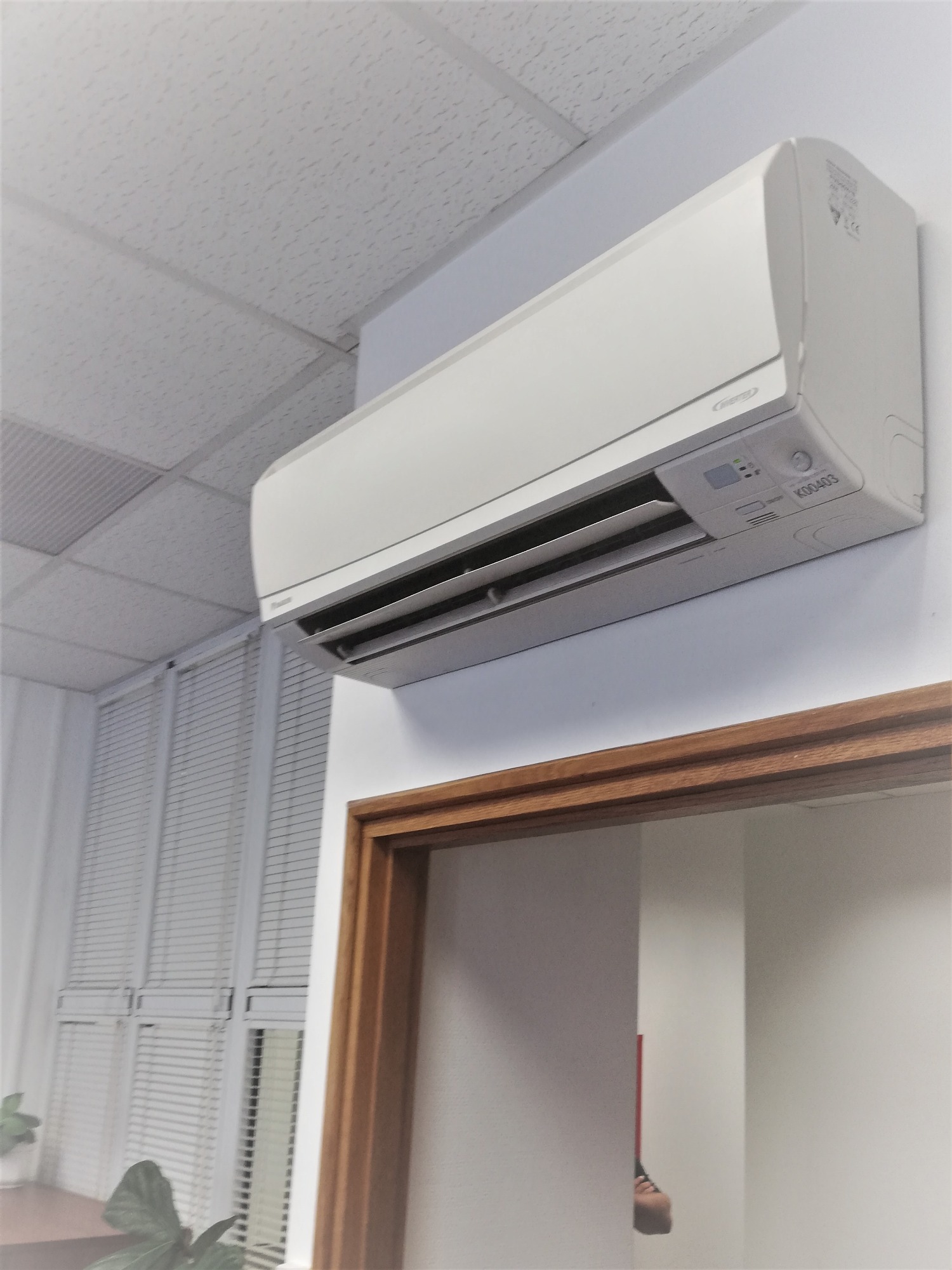 Biały klimatyzator ścienny zamontowany w biurze, widoczny sufit kasetonowy i okno z żaluzjami.