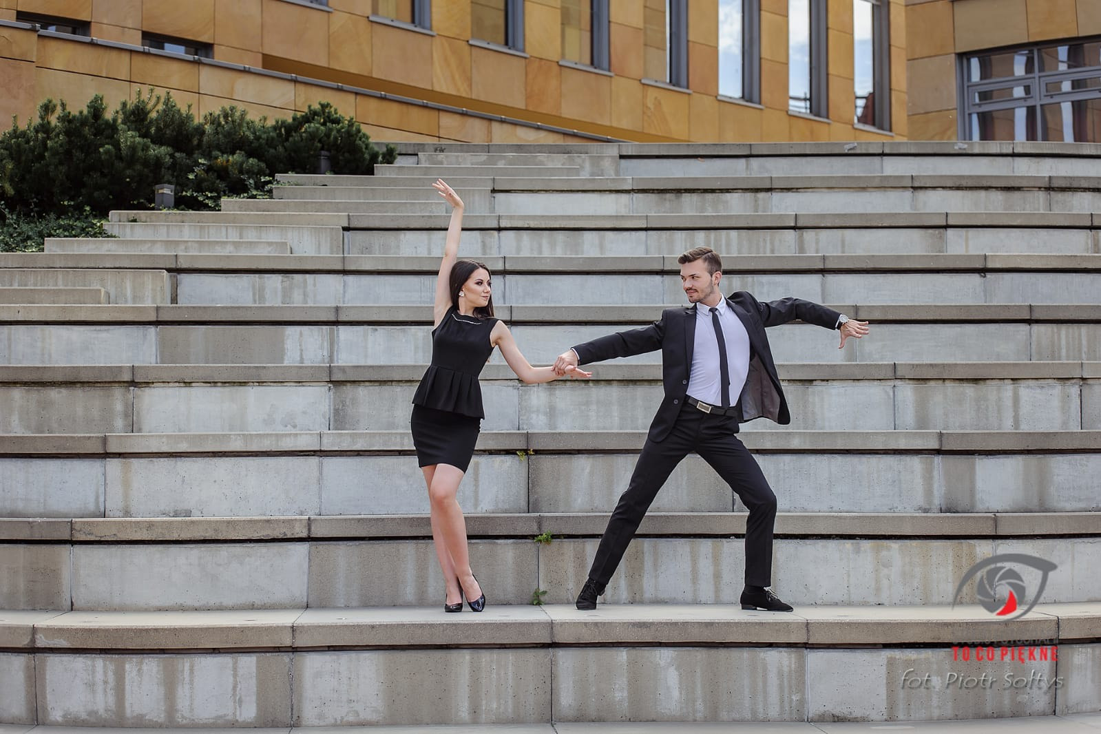Para w czarnych strojach tanecznych pozuje na betonowych schodach przed budynkiem z piaskowca.
