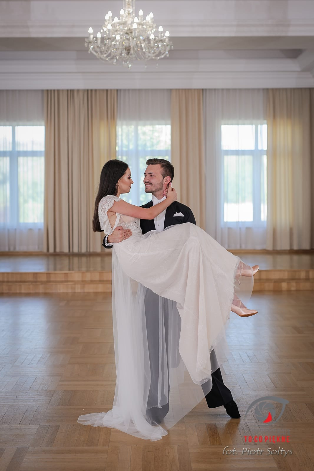 Mężczyzna w smokingu trzyma na rękach kobietę w białej sukni na parkiecie sali balowej z żyrandolem i oknami.