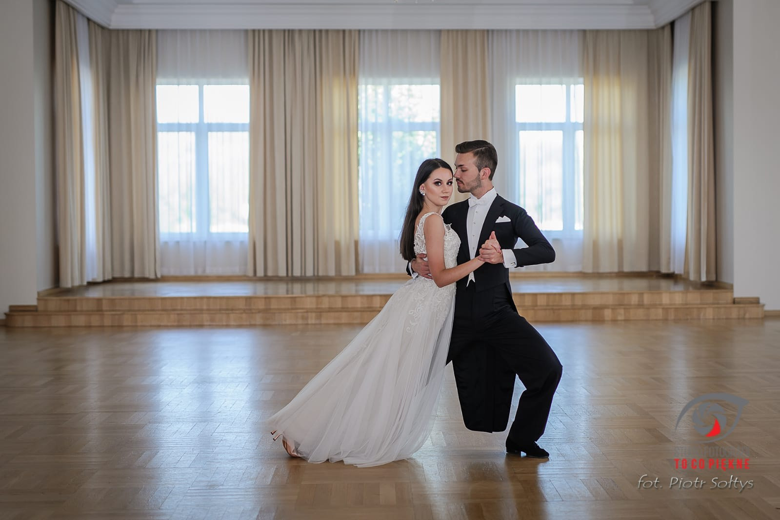 Para w strojach galowych tańczy w przestronnej sali z parkietem i oknami z jasnymi zasłonami.