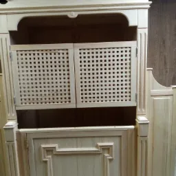 Drewniana szafa w jasnym odcieniu z ażurowymi drzwiczkami, widoczne zdobienia i frezy na elementach konstrukcyjnych.