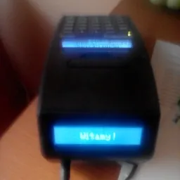 Czarny terminal płatniczy z wyświetlaczem LCD, na którym widnieje napis 'Witamy!' w niebieskiej czcionce, umieszczony na drewnianym blacie.