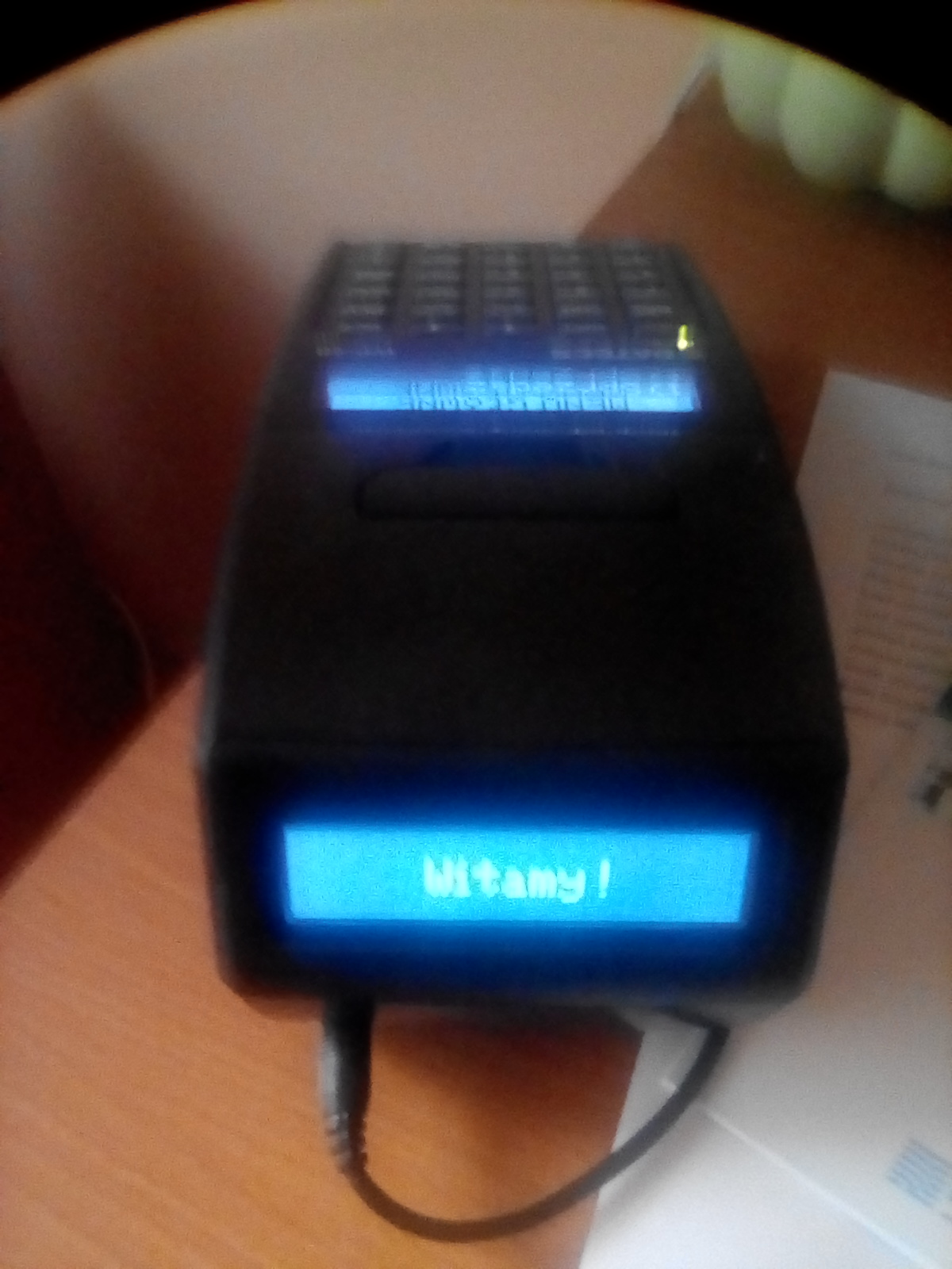 Czarny terminal płatniczy z wyświetlaczem LCD, na którym widnieje napis 'Witamy!' w niebieskiej czcionce, umieszczony na drewnianym blacie.