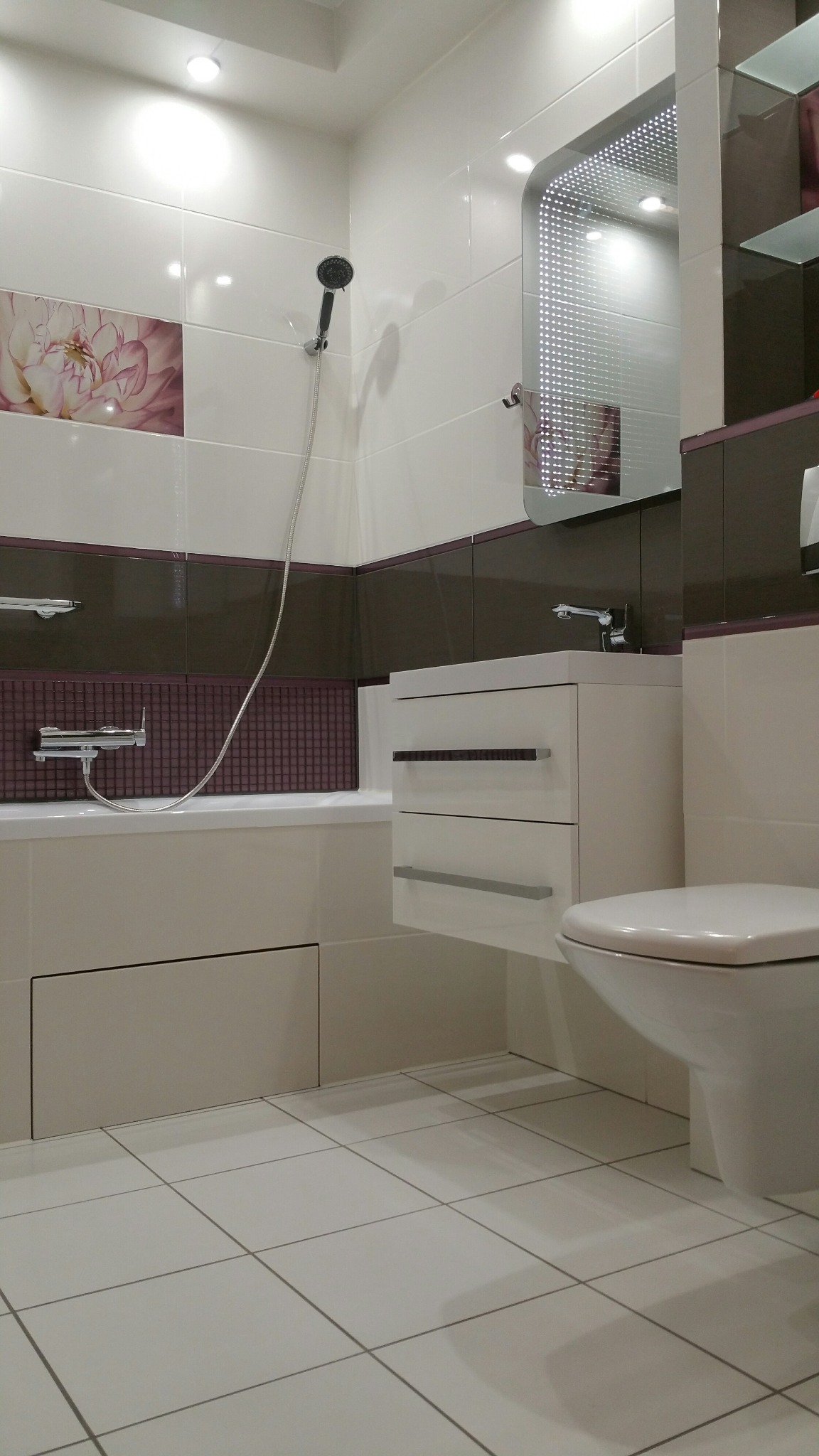 Nowoczesna, wykończona łazienka w Warszawie z białymi i brązowymi płytkami, podświetlanym lustrem, wiszącą szafką z umywalką oraz zabudowaną wanną z prysznicem.