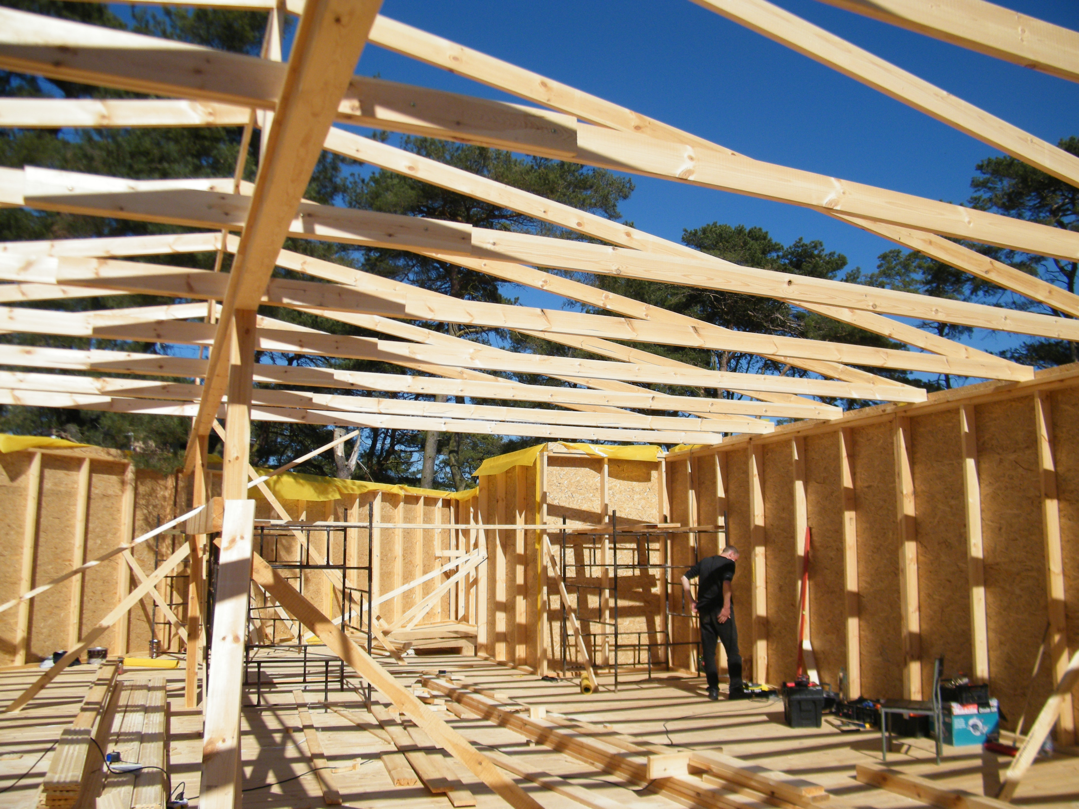 Wnętrze budynku w trakcie budowy szkieletowej z widoczną konstrukcją drewnianą ścian i stropu, na tle błękitnego nieba i zieleni drzew, z pracownikiem porządkującym narzędzia.