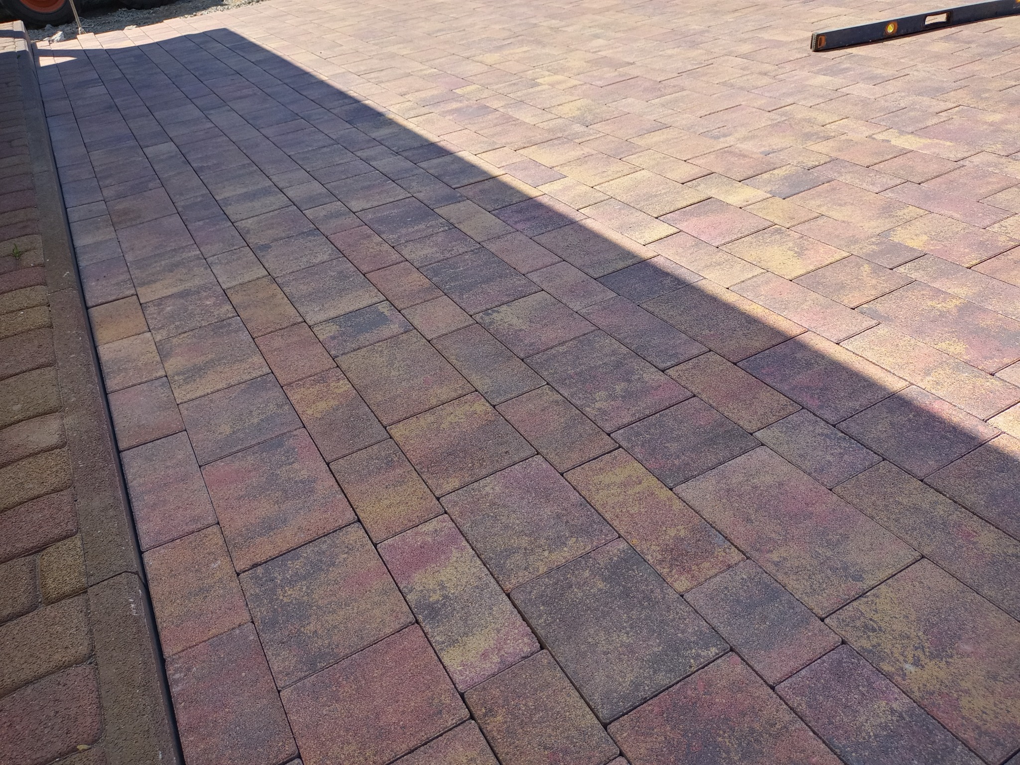 Ułożona kostka brukowa w odcieniach brązu, żółci i czerwieni, oświetlona słońcem z wyraźnym cieniem, z poziomnicą leżącą na kostce w prawym górnym rogu.